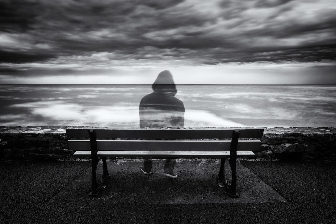 Semi transparent person sits on a bench overlooking ocean in black and white scale image