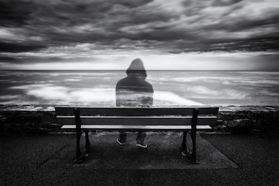 Semi transparent person sits on a bench overlooking ocean in black and white scale image