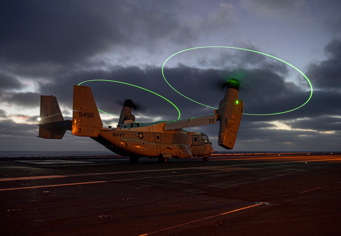 Rotator blades illuminated by green lights spin on a Navy aircraft as it sits on a ship’s flight deck under a dark sky.