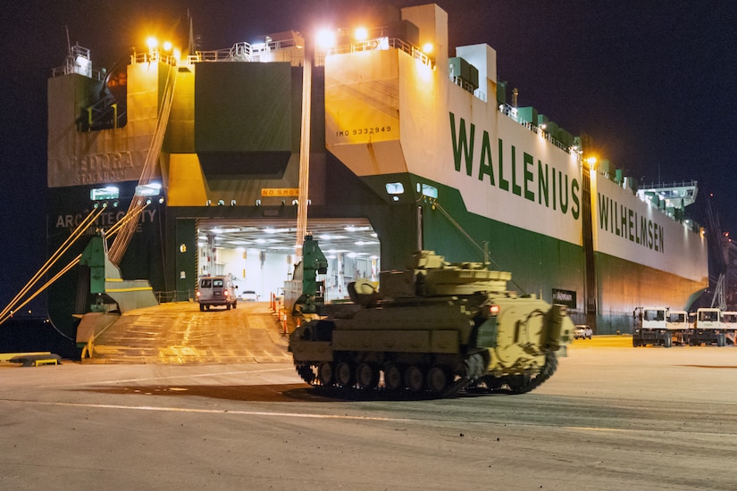A tank sits on a dock near a ship with an open loading ramp.