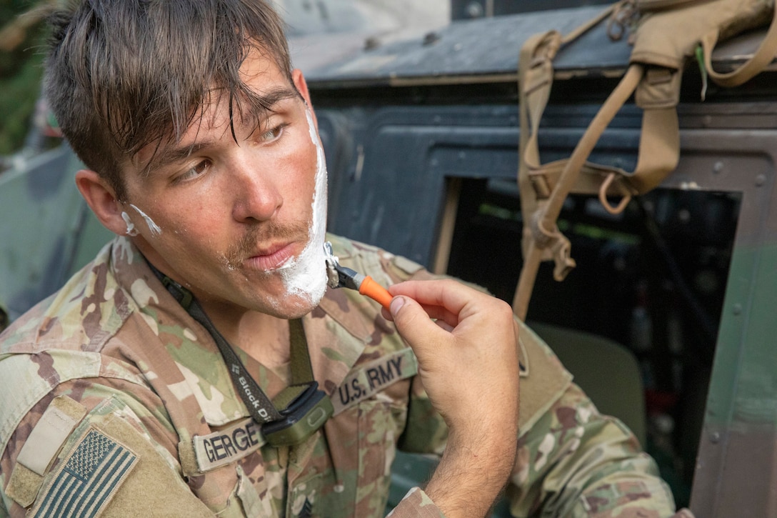 A soldier tilts while using a razor to shave next to a vehicle in a field environment.