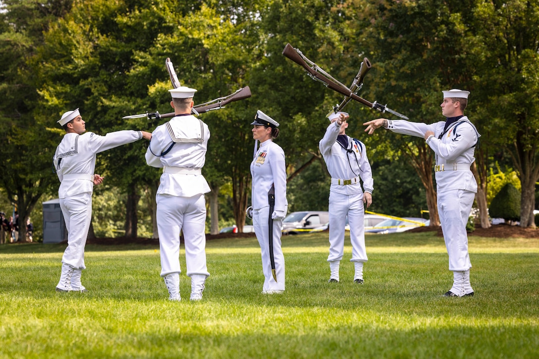 Four members of the Navy Drill Team stand in a square formation while tossing rifles to one another as a fellow member stands in the center and watches.