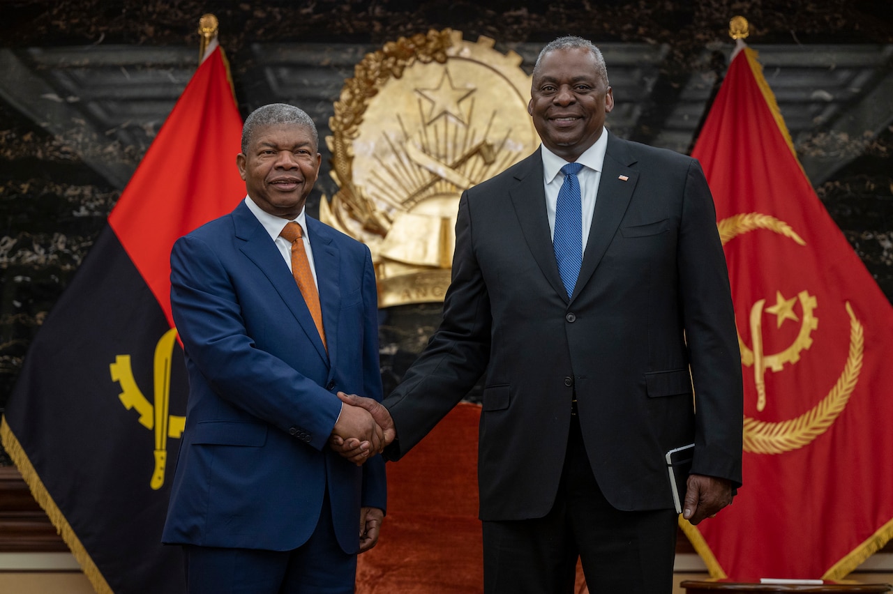 Two men shake hands while standing in front of two flags.