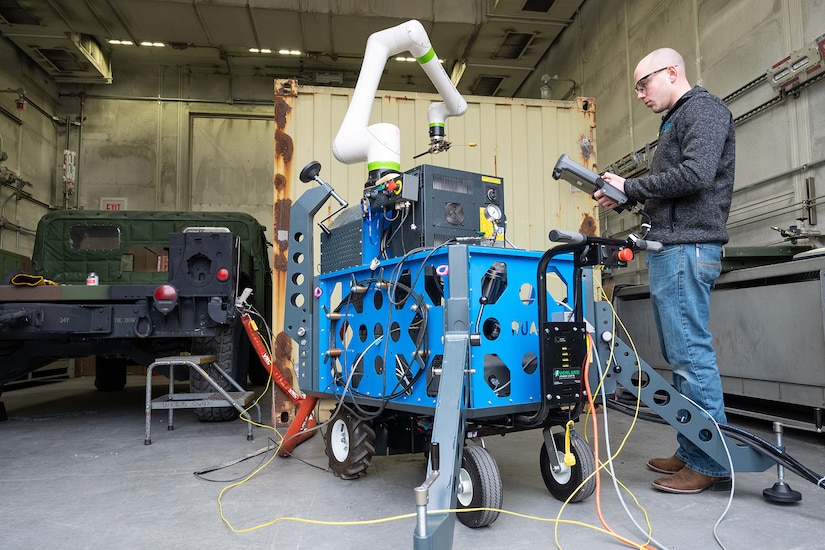 A man in civilian attire stands next to a robotic arm.