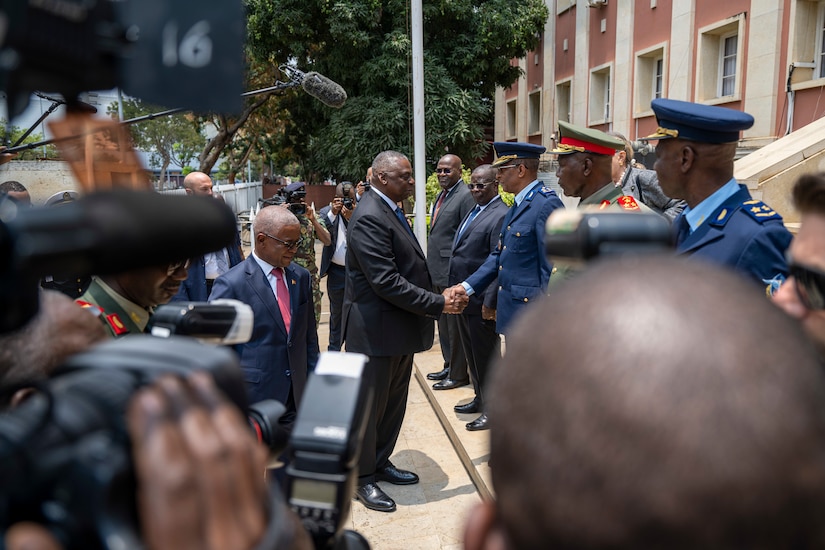 A person in a business suit shakes hands with a person in military uniform as other people watch.