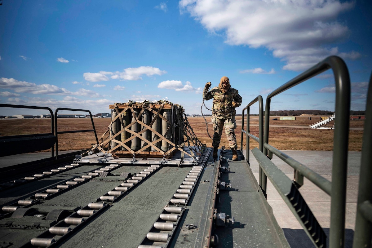 A uniformed service member stands on a flatbed next to a pallet of material.
