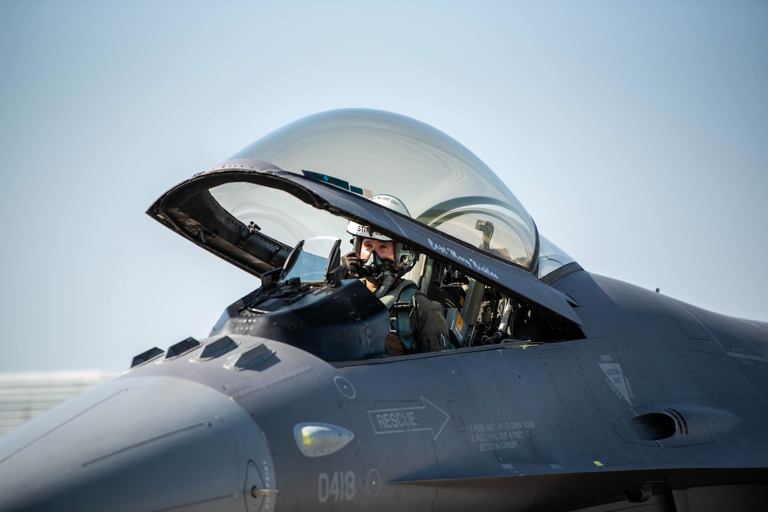 A pilot adjusts his helmet while sitting in a F-16 Fighting Falcon with open canopy.