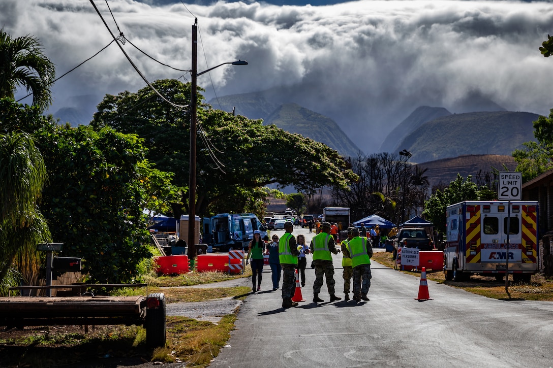 Troops in neon vests stand in a street near roadblocks, with mountains in the background.