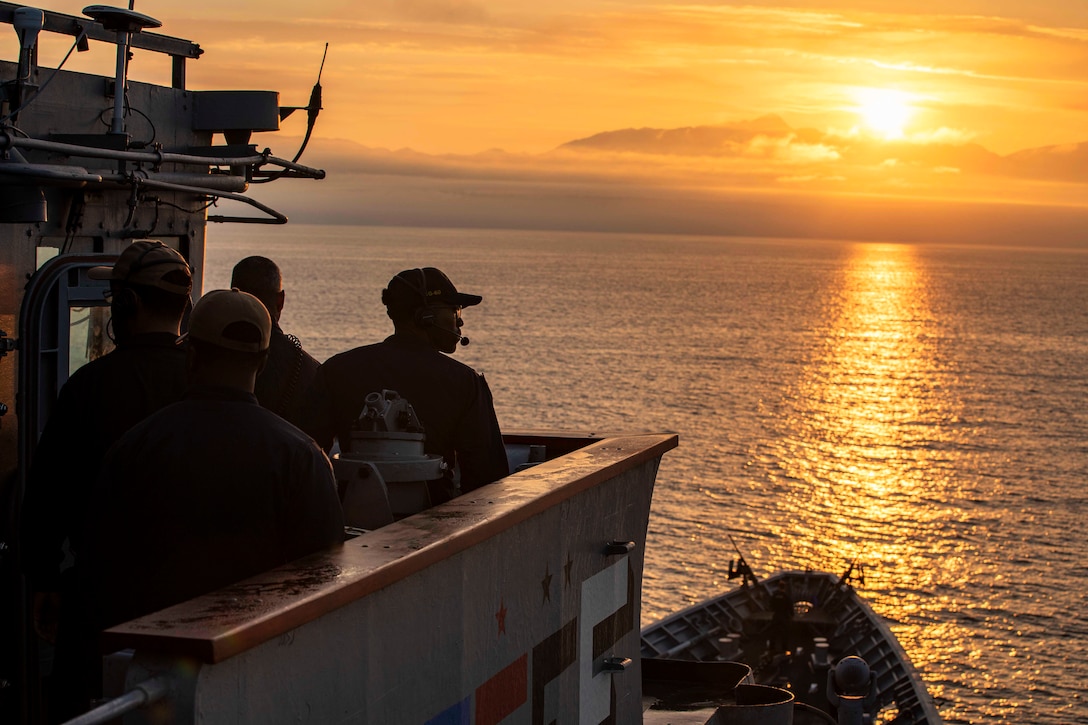 Sailors stand on a ship's deck looking out over water and a low sun in orange sky.