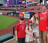 Man in St. Louis Cardinals jersey, woman in a St. Louis Cardinals baseball jersey, young girl in St. Louis Cardinals jersey, and young boy in St. Louis Cardinals t-shirt stand together in front of a baseball field.