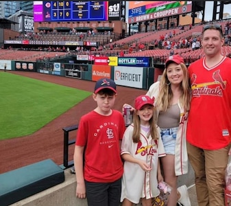 Man in St. Louis Cardinals jersey, woman in a St. Louis Cardinals baseball jersey, young girl in St. Louis Cardinals jersey, and young boy in St. Louis Cardinals t-shirt stand together in front of a baseball field.