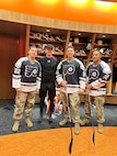 Four men are standing inside a hockey locker room. Three men are wearing Philadelphia Flyers jersey and one man is wearing the undergear for a hockey player.