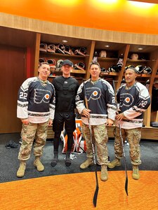 Four men are standing inside a hockey locker room. Three men are wearing Philadelphia Flyers jersey and one man is wearing the undergear for a hockey player.