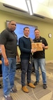 Three men standing in a classroom, man in sweater is being presented an award by a man in a U.S. Army polo