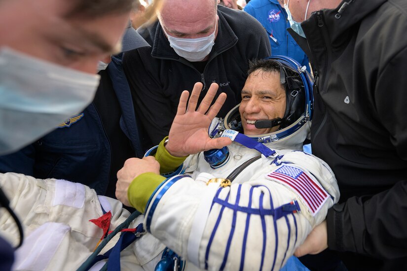 An astronaut waves as they are carried by a few people wearing masks.