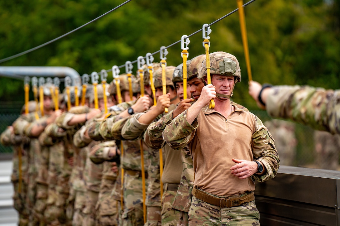 Uniformed service members stand in a line while holding ropes attached to carabiners.