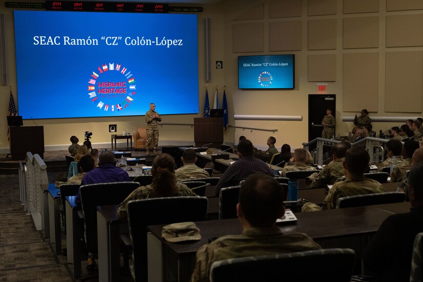 An Air Force military member speaking on a stage to a large group of audience members.