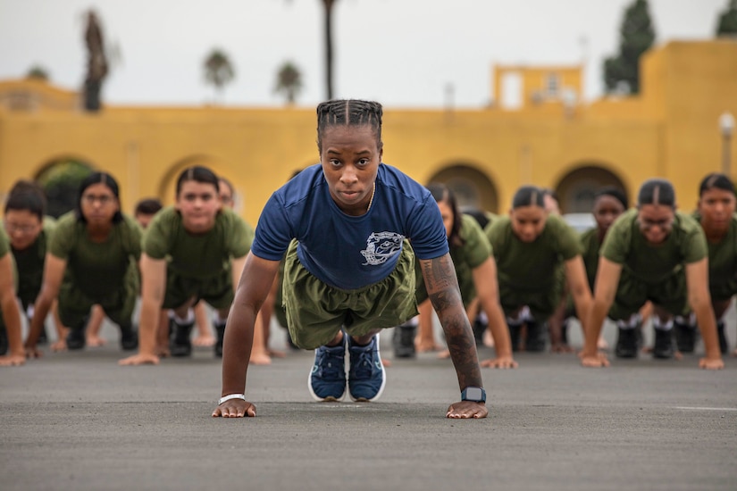 A Marine performs a pushup in front of dozens of fellow Marines also in a push position in a blurred background.