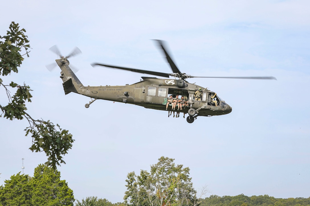 Soldiers sit in the open doorway of a helicopter as it flies over trees.