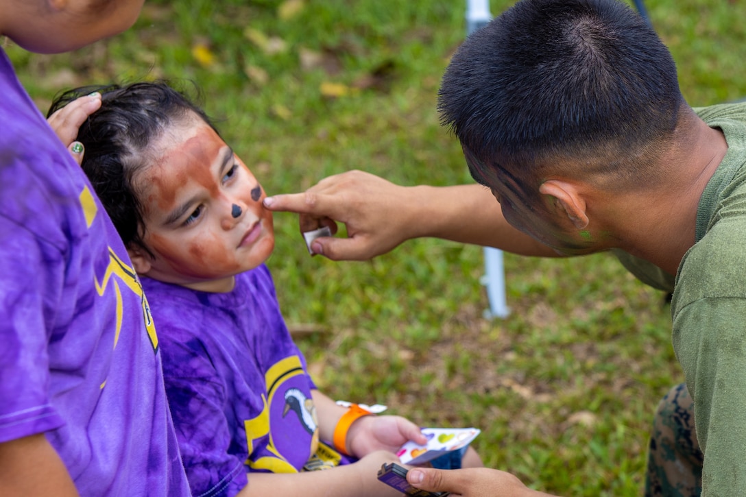 A Marine kneels to apply black polka dots to a child’s face as another child stands nearby.