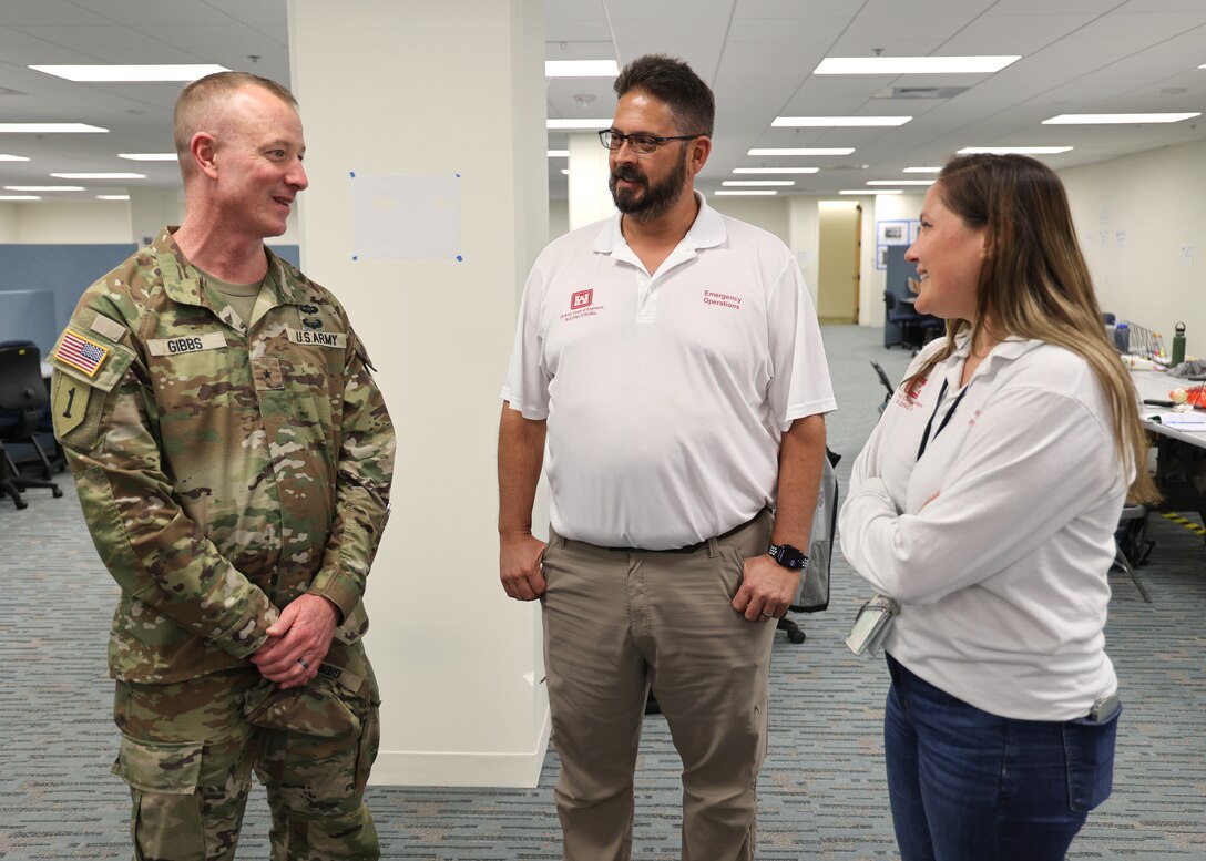 Three people talk in an office setting. One of the people wears the uniform of an Army brigadier general.