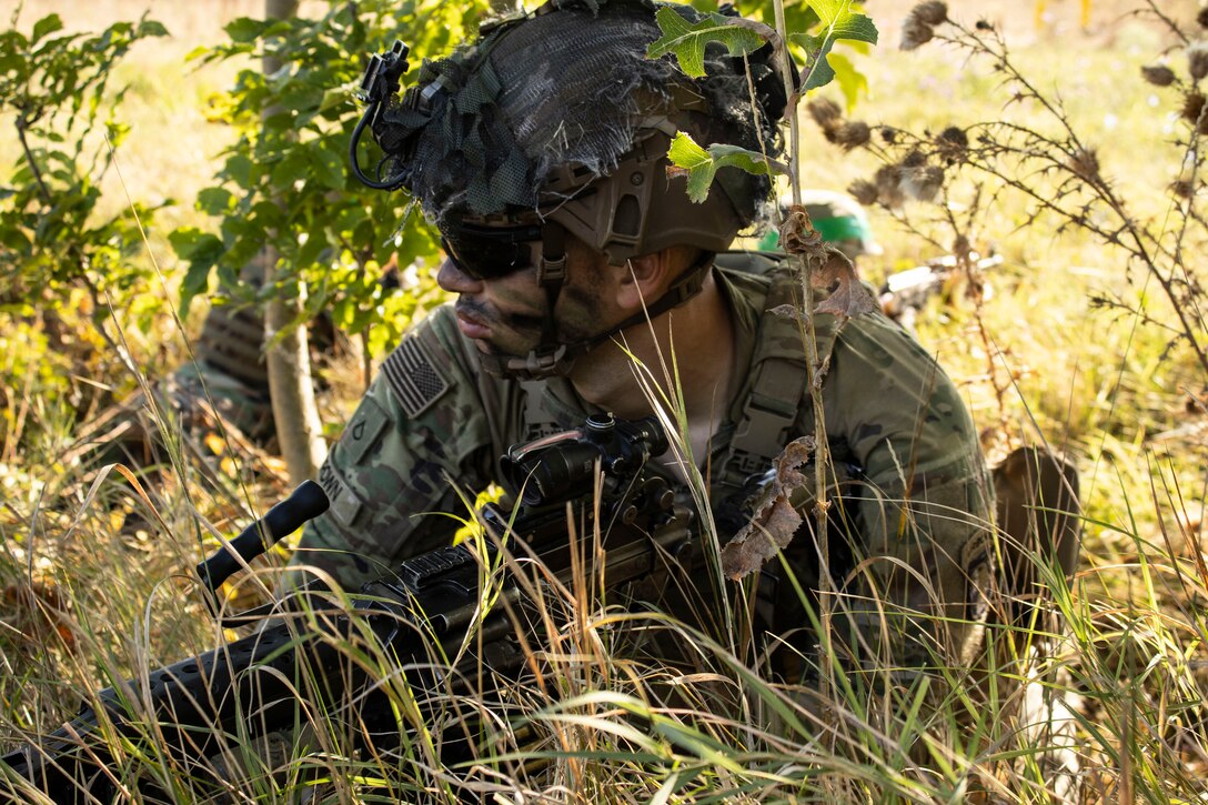 A soldier laying in tall grass takes cover while participating in a field training exercise.