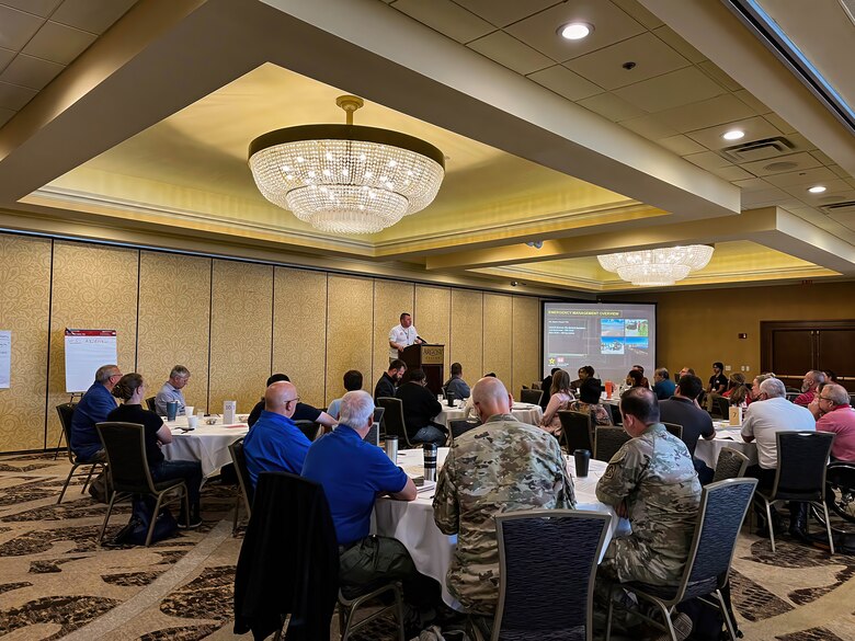 A large room with circular tables with white table cloths and people seated around the tables listen to a speaker at a podium in the background.