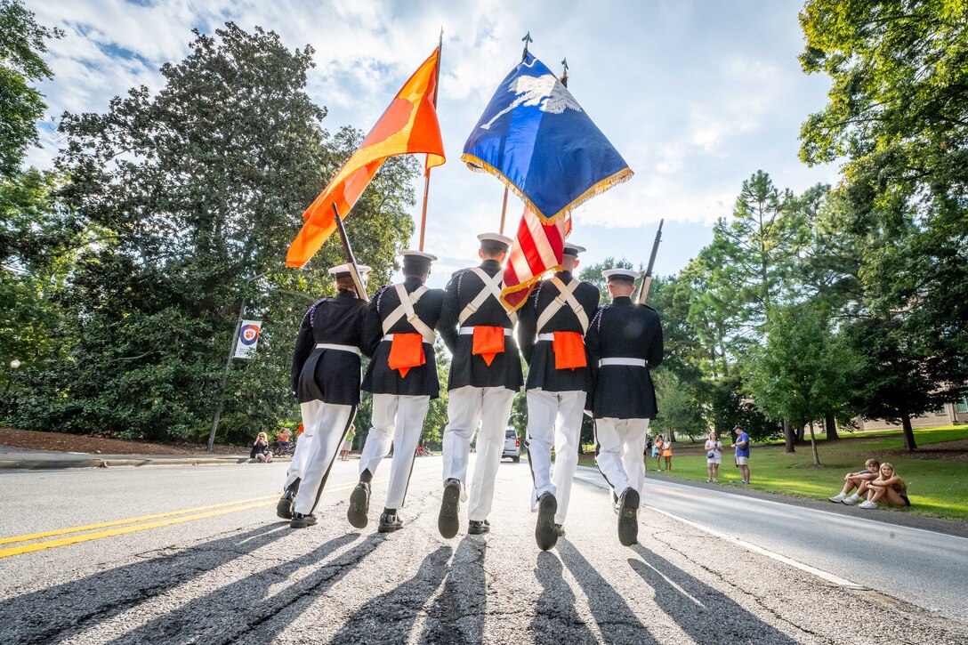 Five Army ROTC cadets holding flags and ceremonial weapons march in a parade. The cadets are photographed from behind.