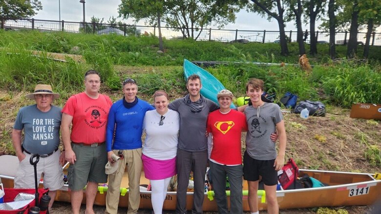 Seven people stand with their arms around each other in front of a kayak with geen grass and trees in the background.
