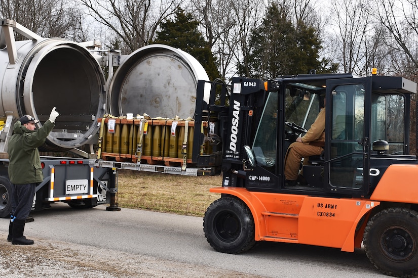 A person in civilian attire directs a forklift driver moving a pallet of artillery rounds onto a truck trailer.