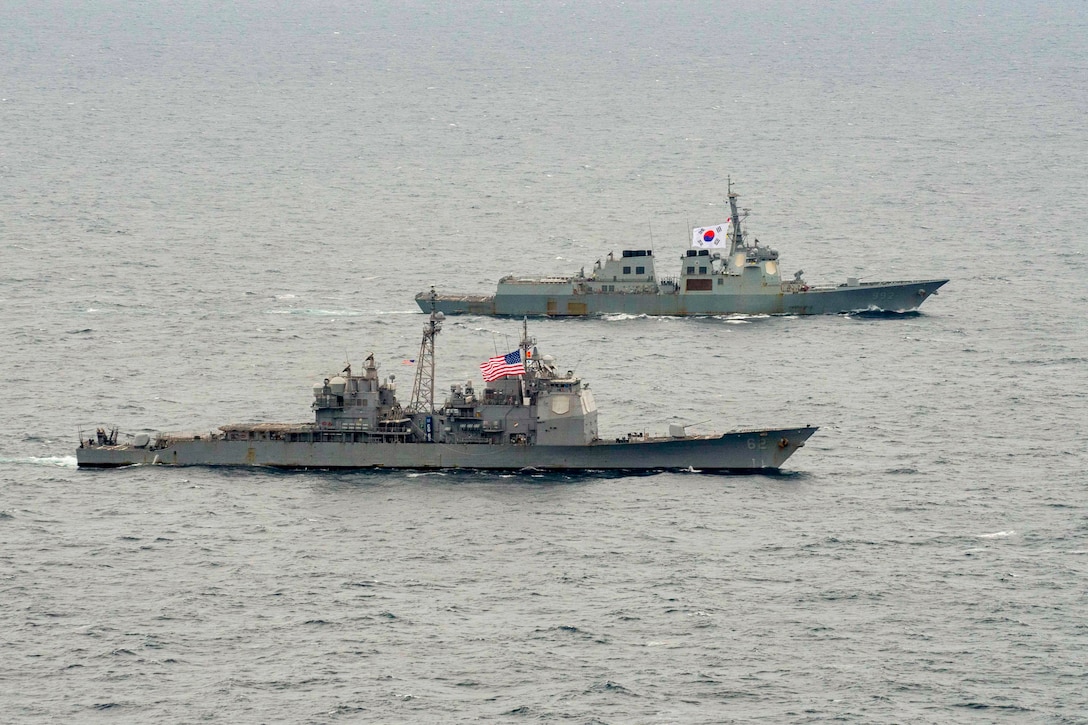 One ship displays an American flag as another displays a South Korean flag while they sail in formation in a body of water.