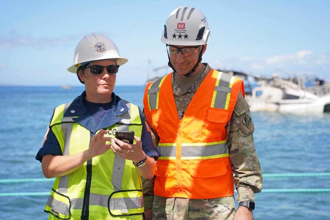 A man and woman look at a smartphone. The pair are wearing safety gear and hardhats. The ocean is visible behind them including a damaged vessel.