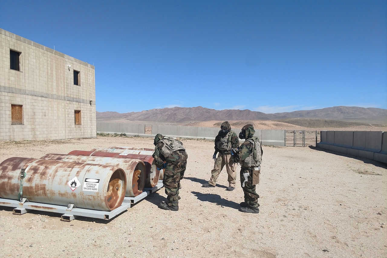 People wearing protective suits stand near metal cylinders.