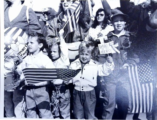Young children and family members wave United States of America flags and signs, welcoming home service members.