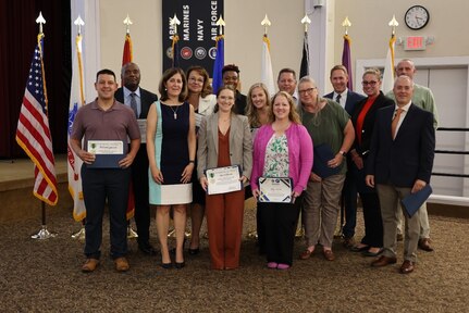 A group of 15 staff stand with awards and official US flags are in the background