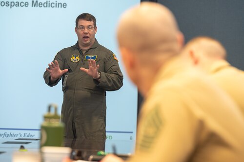 An Air Force medical officer stands at the front of the room, while two U.S. Marine Corps Sergeant Majors can be seen watching him.