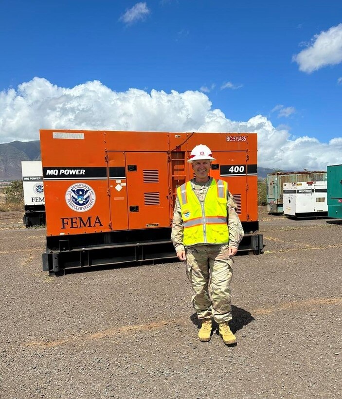 A man in a military uniform, hardhat and safety vest stands in front of an industrial power generator.