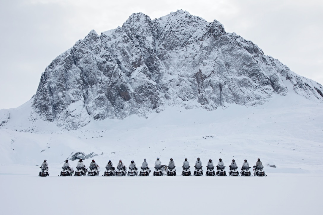 A group of service members in winter gear pose for a photo sitting on snowmobiles with a mountain in the background.