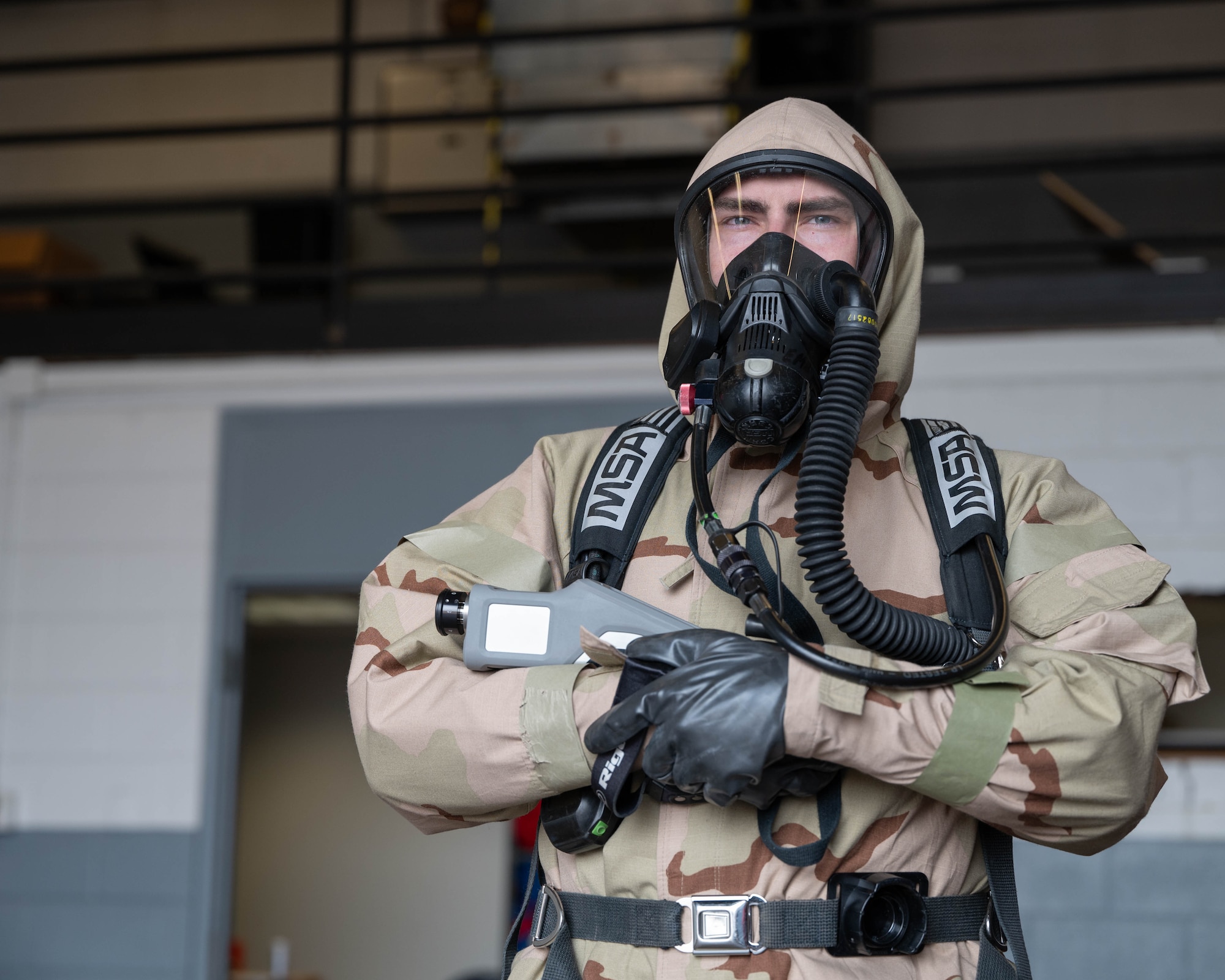 Senior Airman Ruehle is wearing mission-oriented protective posture gear and the self-contained breathing apparatus while holding the Raman spectrometer.