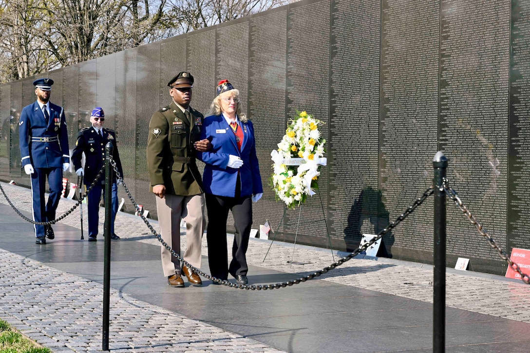 Two service members walk next to two veterans with a memorial wall and wreath to the right and trees in the background.