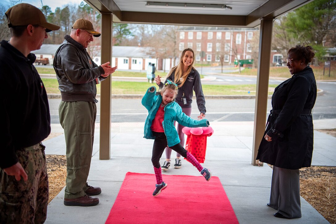 A child dances on a red carpet at a building entryway with an adult walking behind. Three adults flanking the carpet look on; one claps.
