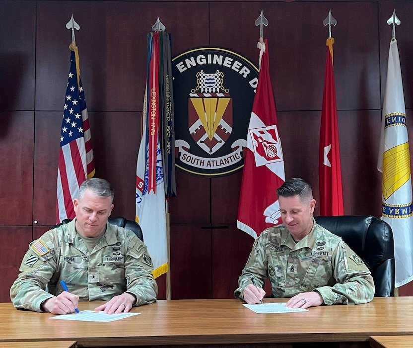 2 Army men in camouflage uniforms signing documents in front of U.S. and Army flags.