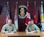 2 Army men in camouflage uniforms signing documents in front of U.S. and Army flags.