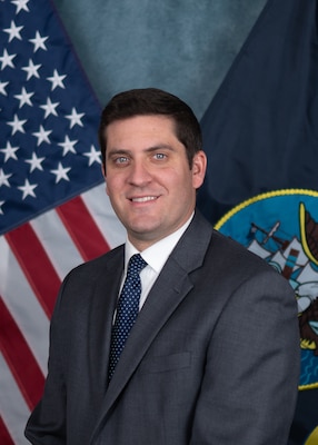 A photo in front of the U.S. flag and the U.S. Navy flag of David Macht, the director for U.S. Navy Strategic Systems Programs (SSP) Mission Support. David is smiling and has on a slate gray suit with a dark blue tie and a white collared shirt.