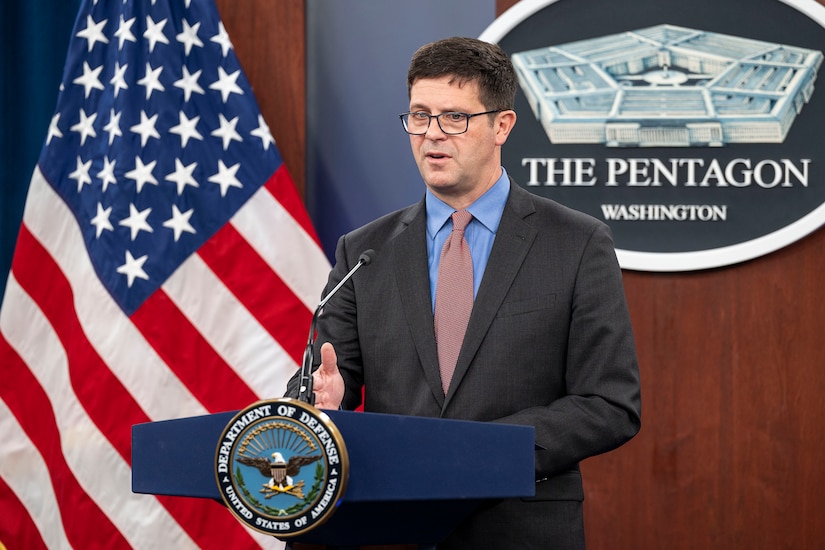 A civilian stands and speaks into a microphone at a lectern, with a U.S. flag in the background.
