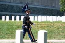 Military members who are wearing dark Army ceremonial uniforms are marching down a road with rows of identical white tombstones in the background and foreground
