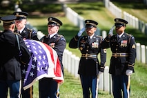 Two men in dark Army ceremonial uniforms are saluting towards other soldiers who are carrying a casket that is draped with the US flag. There are rows of white tombstones and green grass in the background.