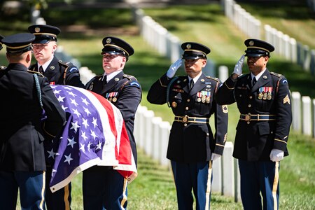 Two men in dark Army ceremonial uniforms are saluting towards other soldiers who are carrying a casket that is draped with the US flag. There are rows of white tombstones and green grass in the background.