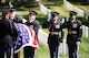 Two men in dark Army ceremonial uniforms are saluting towards other soldiers who are carrying a casket that is draped with the US flag. There are rows of white tombstones and green grass in the background.