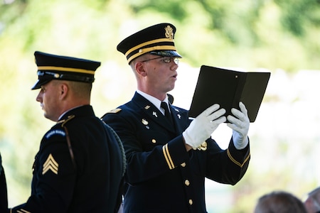 An Army soldier in a dark ceremonial unform and white gloves is holding a black book while reading.